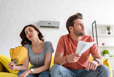 Two people sitting on a couch fanning themselves because their outside AC is not working.